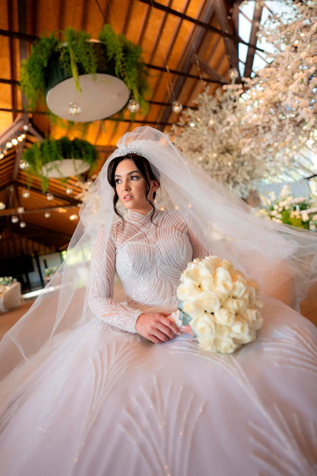 Noiva em pé com buquê branco diante de arranjo floral, com véu longo