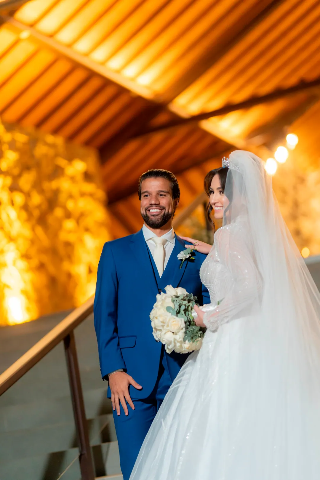 Casal de noivos no altar, sob lustre de cristal — noivo de terno e noiva de vestido bordado com véu longo
