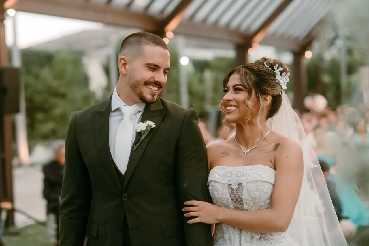 Noivos abraçados sob arco de flores brancas e lustre de cristal, casamento em estrutura de vidro